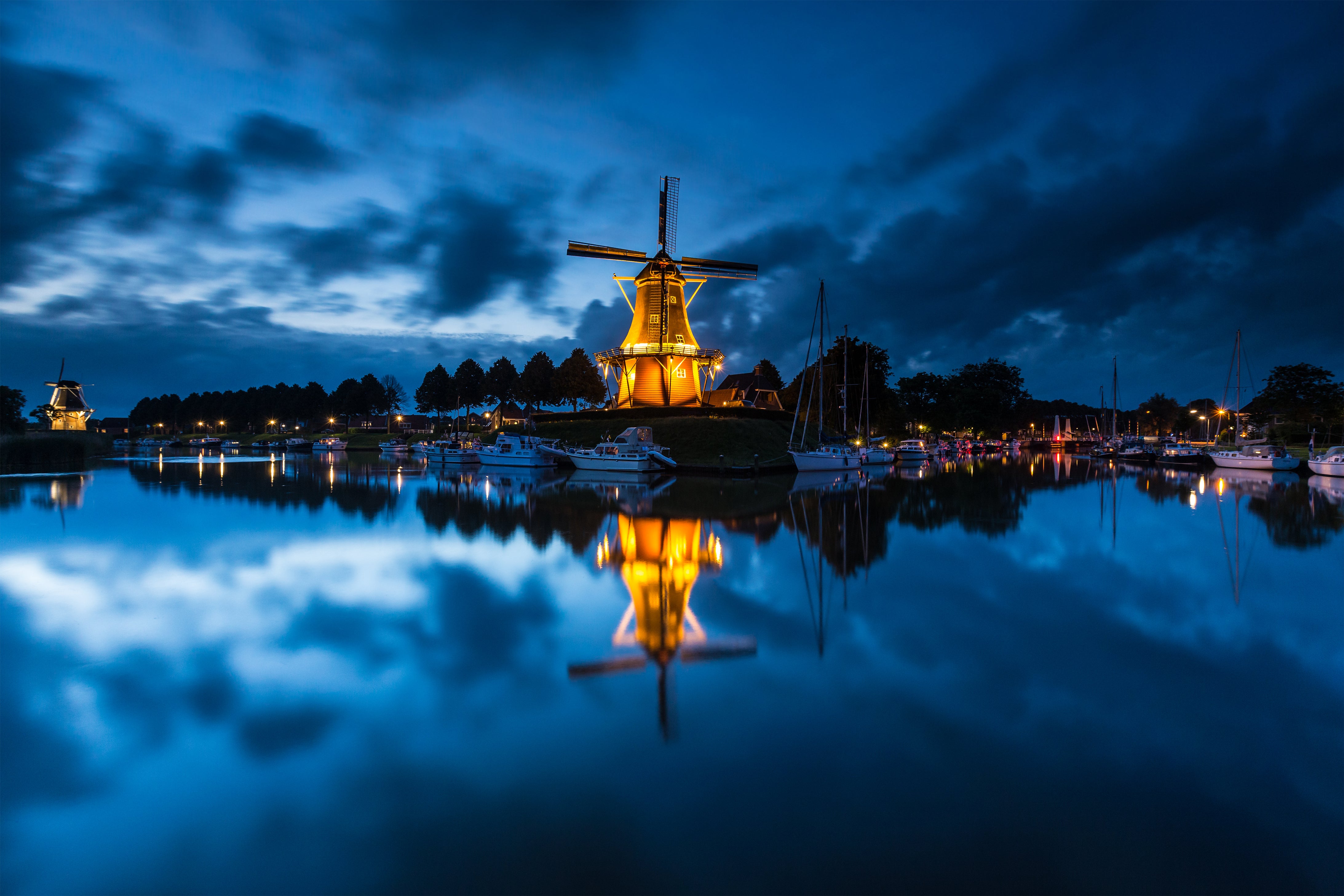 Illuminated windmill reflecting in water at night with boats and trees in the background