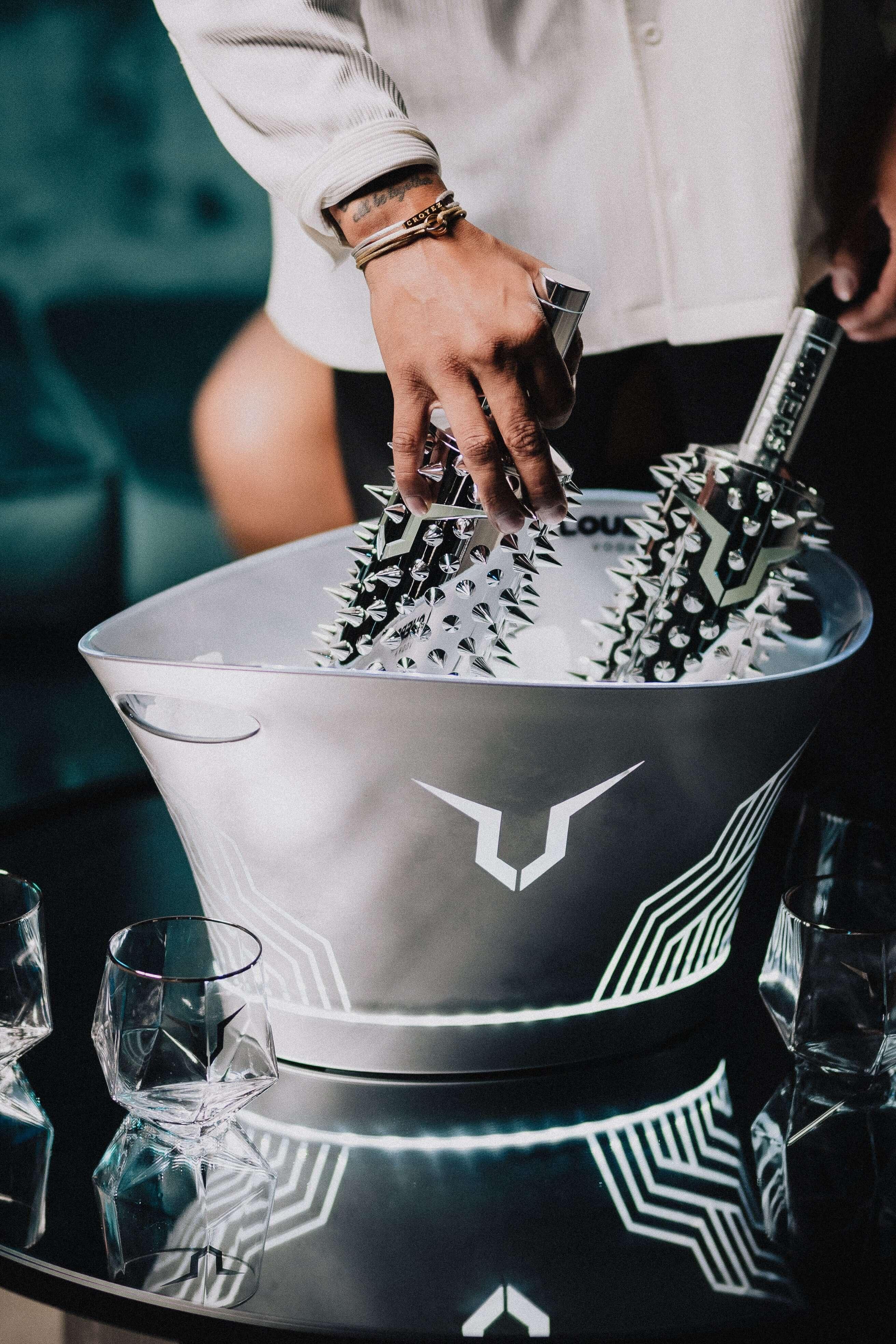 Hand placing luxury bottles in an illuminated ice bucket on a glass table with geometric glasses.