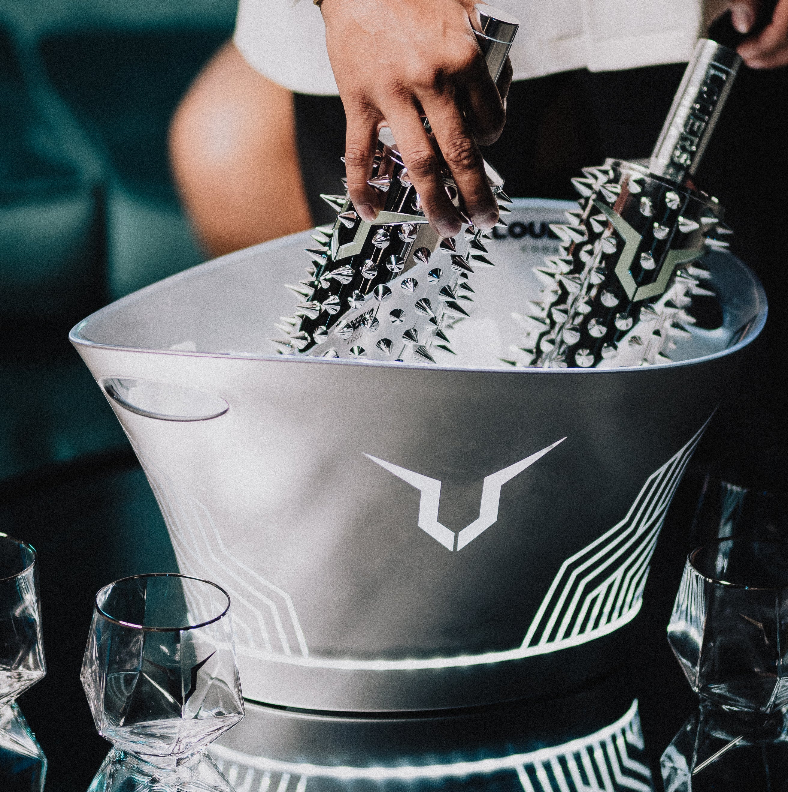 Person placing spiked metal objects into a silver ice bucket with geometric design, surrounded by glasses on a table.